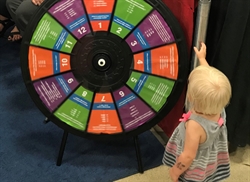 [ai] A young child with light blonde hair reaches towards a colorful spinning wheel marked with numbers and sections in bright colors. The wheel is set against a backdrop of black fabric and visible attendees are in the background.